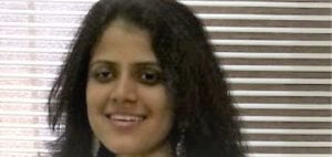 Headshot of a smiling young female graduate student with dark hair and eyes in front of a window with blinds.