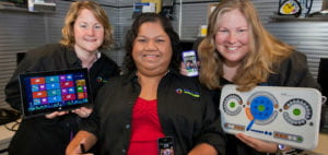 A woman on the left holds a Microsoft Surface tablet. A woman in the center holds a smartphone. A woman on the right holds a smartphone in her right hand and a modified keyboard with her left hand.