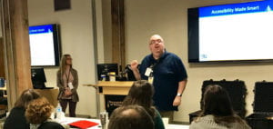 A woman and a man stand in front of a podium and 2 LCD screens in a room of workshop participants. 