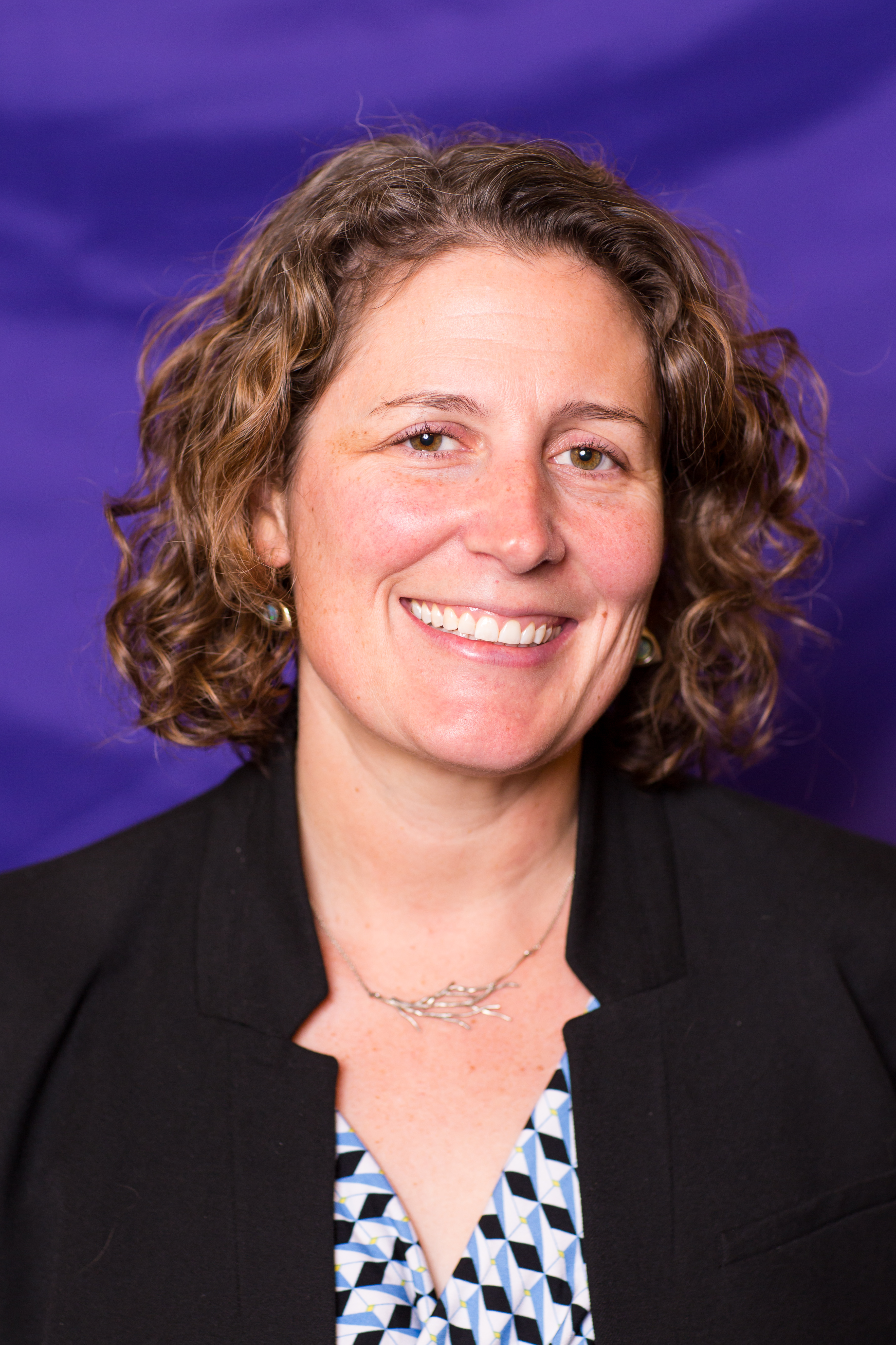 photo of a white woman with curly, light-brown hair, smiling in front of a purple backdrop 