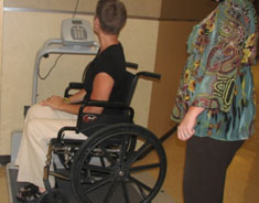 A wheel chair user looks at a digital readout while chair rests on a large black platform. Another person stands nearby