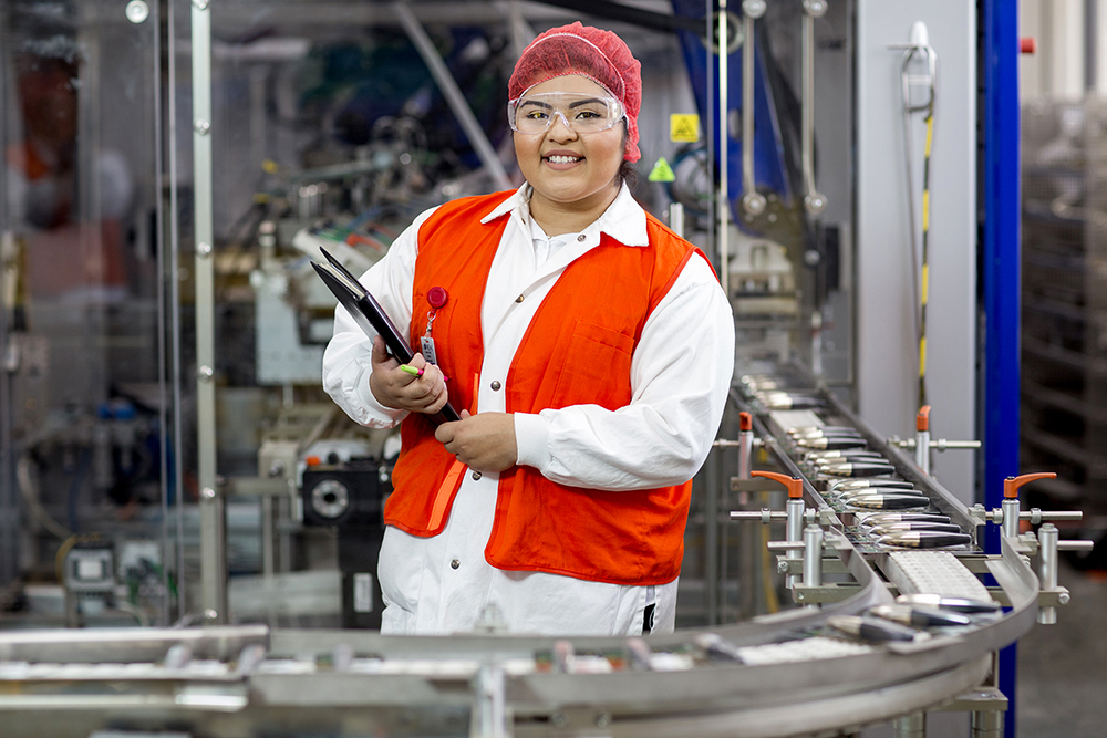 Female student in safety gear, holding clipboard, in a factory setting. 