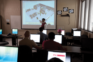 students in a modern-day classroom watch a professor pointing out something on a screen