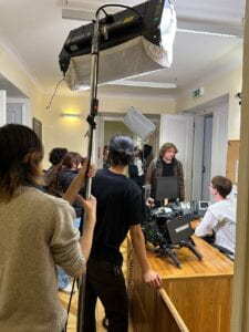 Film students crowd around the NYU academic center's reception desk in preparation to shoot a scene. They hold camera and lighting rigs