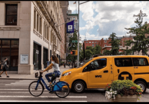 student on bike rides in front of NYU building as nyc taxi passes