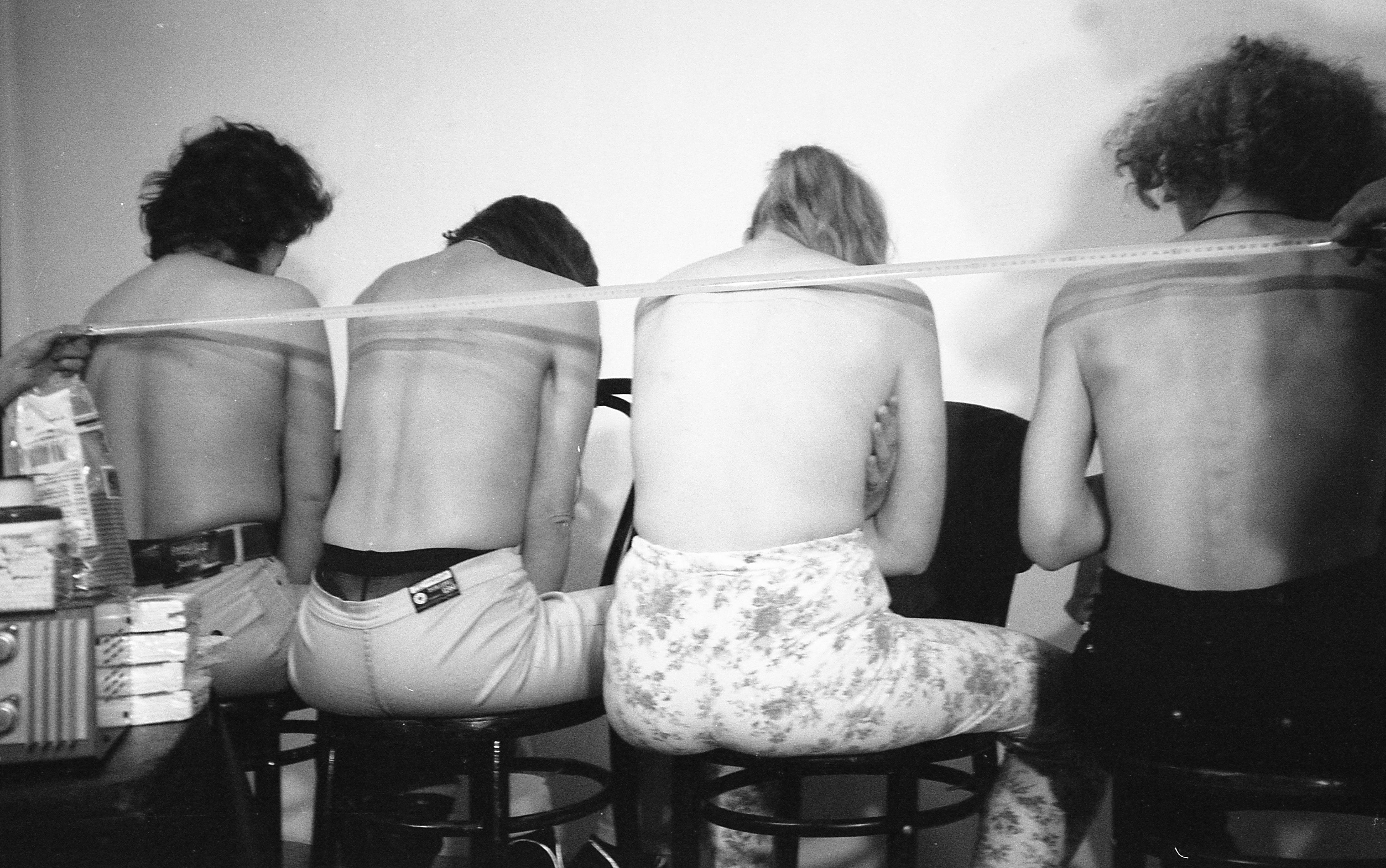 A black and white still image from Sierra’s performance video shows four topless women seated with their backs facing the viewer. A long tape measure is being held across their backs in preparation to receive tattoos.