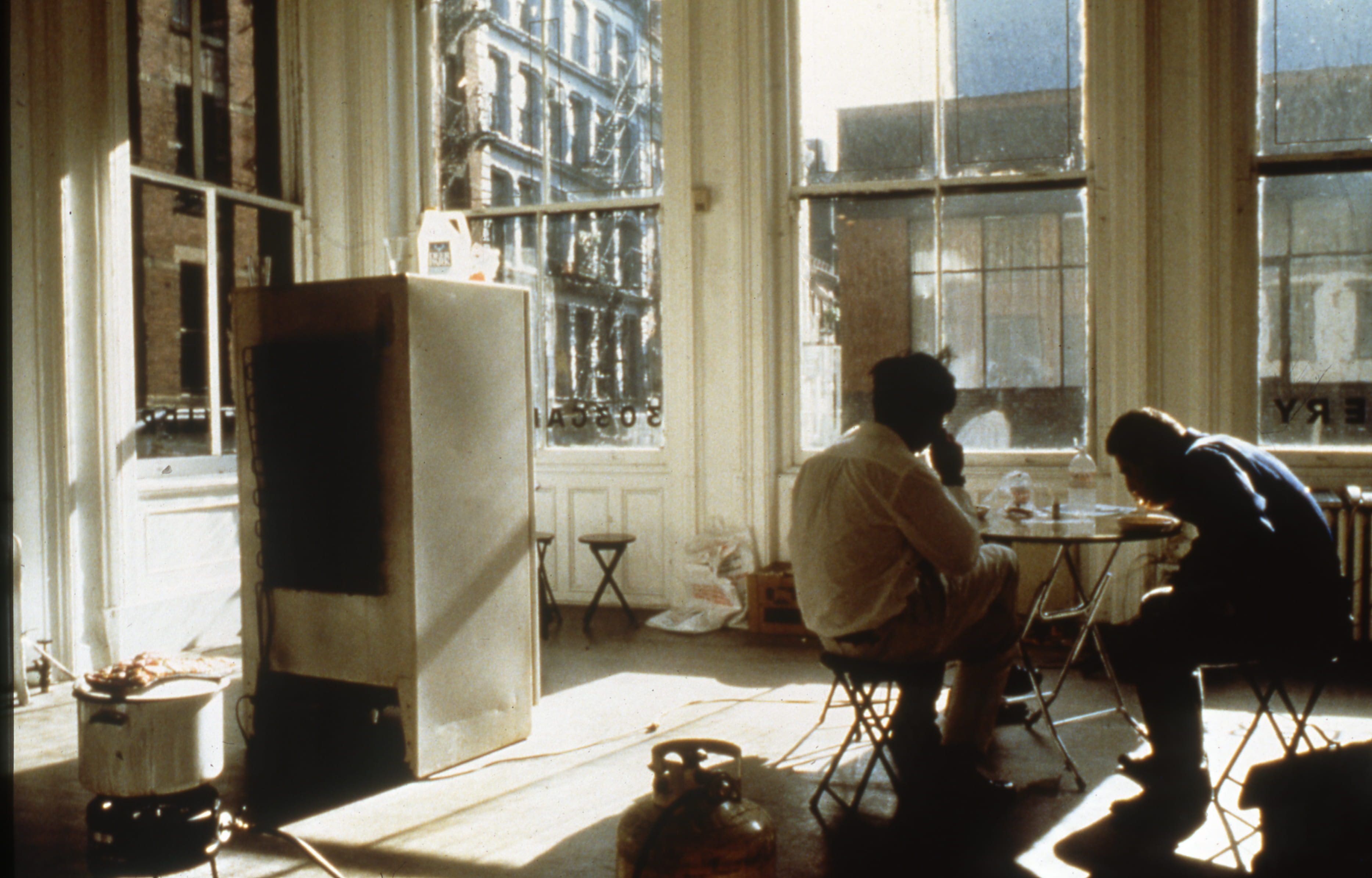 A color photograph from Tiravanija's open house shows two men eating rice and curry at a small table with folding chairs. The four large windows behind them reveal the surrounding city, and backwards window text indicates they are inside the 303 Gallery. 