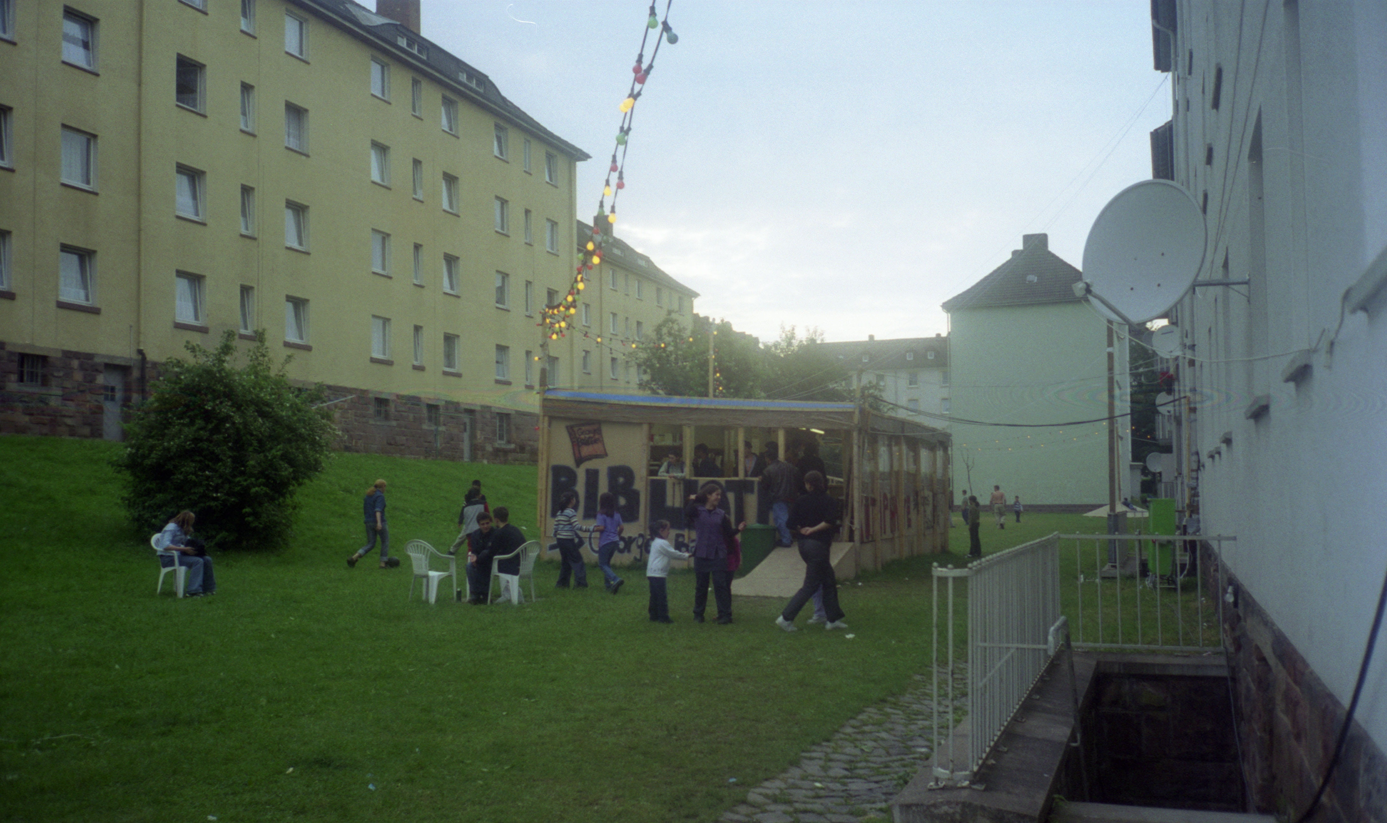 A color photograph shows the constructed library from Hirschhorn’s Bataille Monument in Kassel. The library is built on the grass lawn between two large apartment buildings. Parents and children explore the library; some are sitting in lawn chairs outside of it, while others mill around the interior. Red, yellow, and green lights are strung up above the library giving it a festive mood.