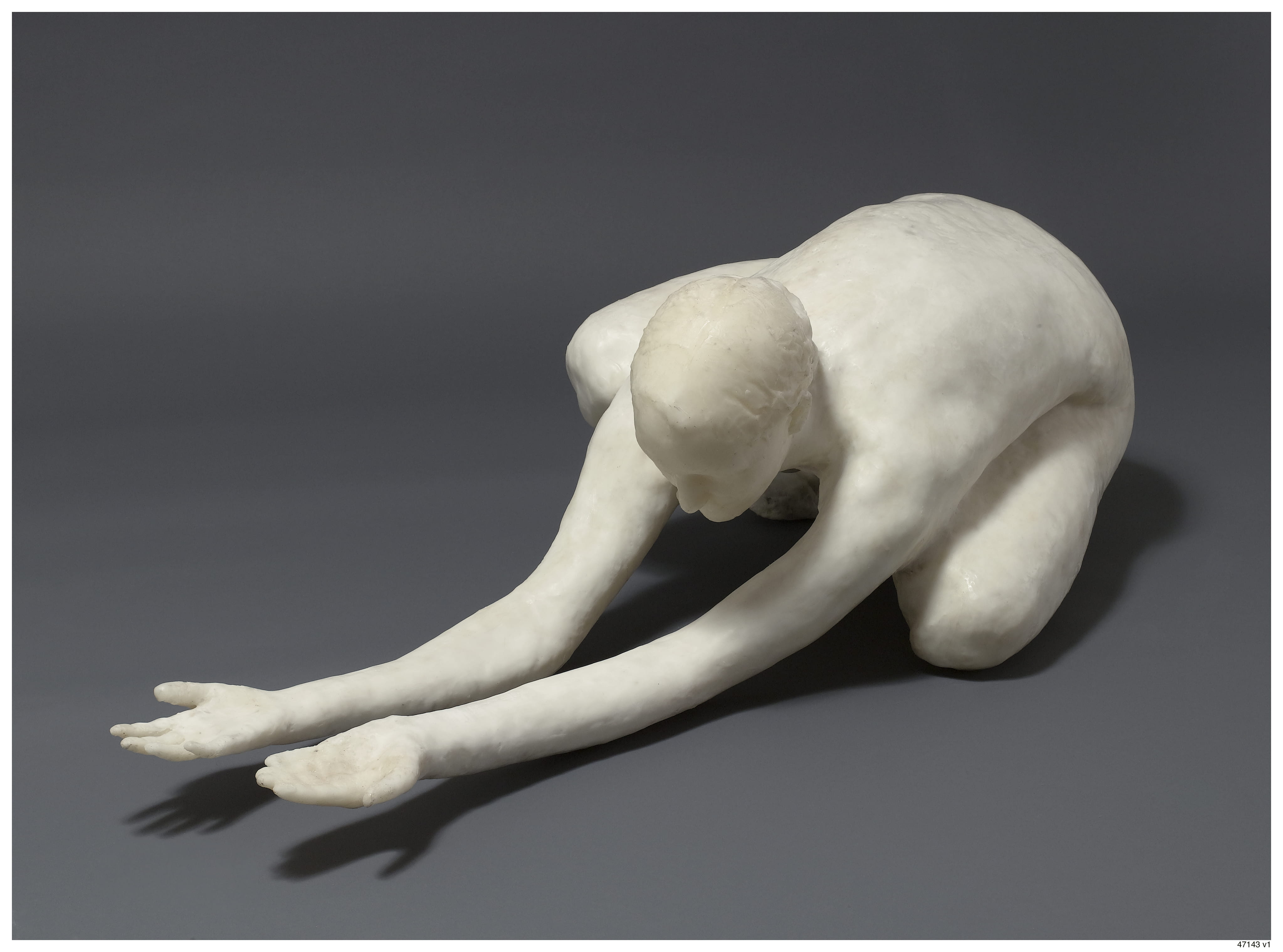 A color photograph shows one of Smith’s sculptures placed on a gray backdrop. The sculpture is white in color and portrays a human form squatting on the ground, legs tucked beneath them. Their torso is bent almost flat against the top of their thighs, and the figure’s arms are fully outstretched in front of them with the palms of their hands facing up. The figure’s gaze does not meet the viewer’s, but instead looks towards the floor near their hands.