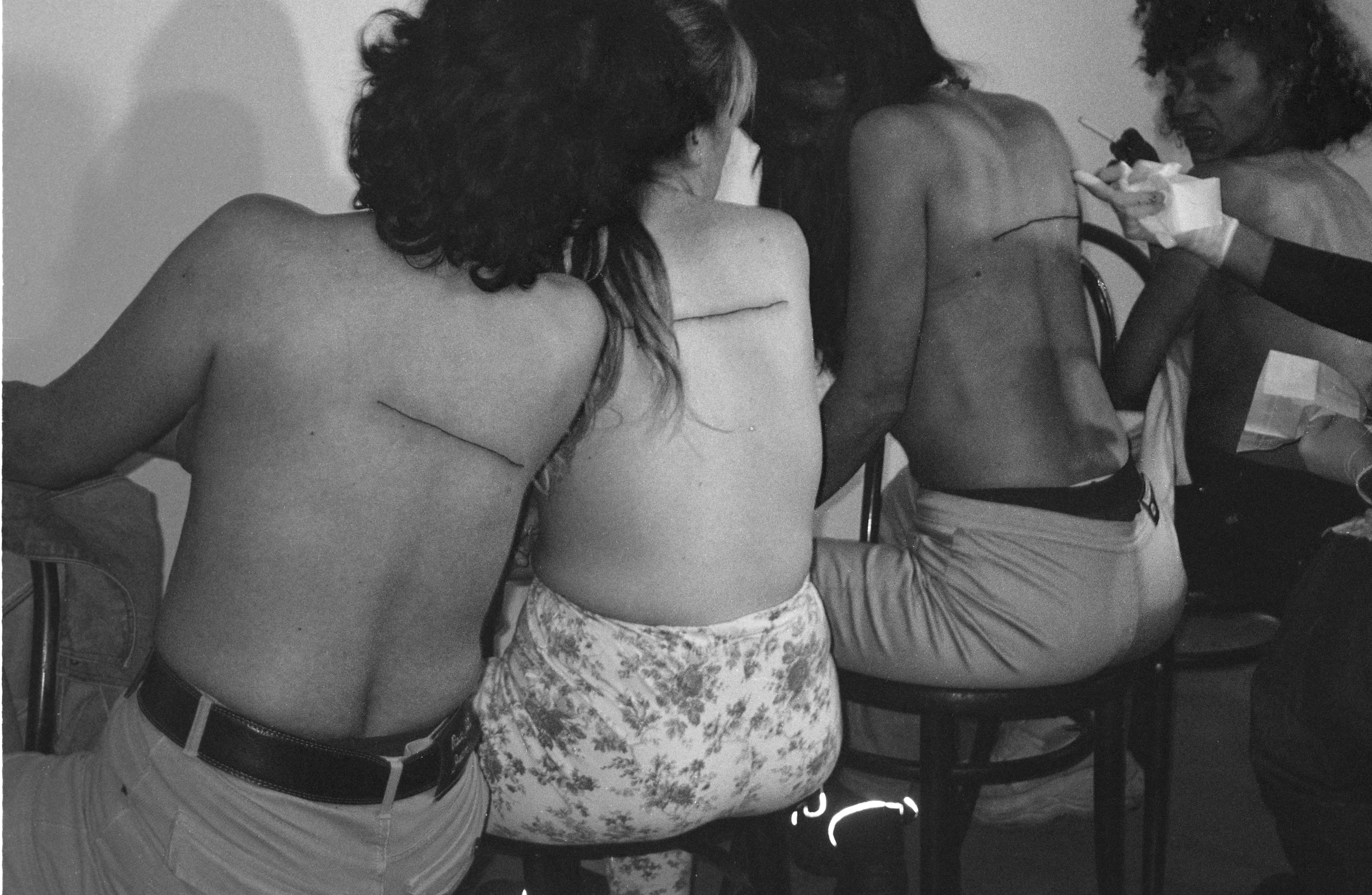 A black and white still image from Sierra’s performance video shows the same four, topless women in the midst of being tattooed. Starting from the left, the first two women already have a tattooed line on their backs. They are both turned to watch as the third women gets tattooed. The last woman on the right also watches, grimacing, in anticipation of her own tattoo. 