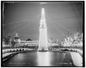 In this aerial view of Coney Island, an isolated tower rises above a calm body of water, surpassing all other structures in height. Its entire architecture sparkles with electric light, illuminating the entire scene, and multiple strings of lights connect its pointed roof to the buildings around it.