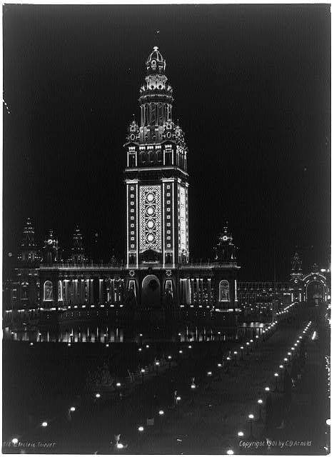 [In this nocturnal cityscape, a large building topped with a tall, ornate tower emerges from the darkness. Electric lights outline the contour of its architectural elements.