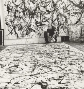 The photograph depicts, in black and white, Jackson Pollock working in his studio. Pollock is situated in the background, towards the righthand edge of the frame. Behind him is a massive canvas on which he has painted work and in front of him, a second canvas covers the floor. He studies it as though assessing its completion.