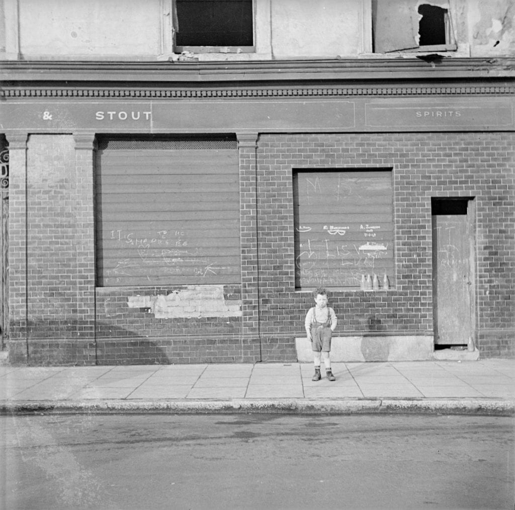 Nigel Henderson's photograph shows a young boy standing solemnly in front of a storefront, likely a bar, with his hands in the pockets of his overalls. The street, in the foreground, and the sidewalk are empty.