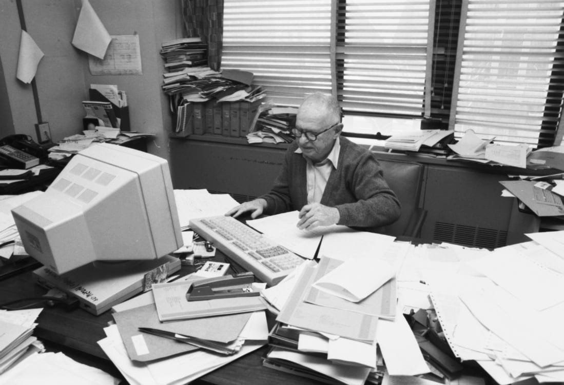 Max Goldstein sitting behind a large computer monitor and a desk covered in dot matrix print-outs