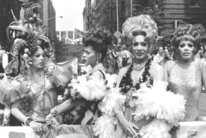 Four persons wearing elaborate headgear or bouffant hairdos, jewelry, and evening gowns carry a banner at an daytime outdoor march in New York City. Fales Library. (Details to be added)