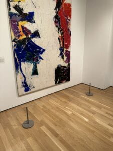 A large colorful painting on a gallery wall; a thin wire barrier stands on the ground a few feet in front of the painting.