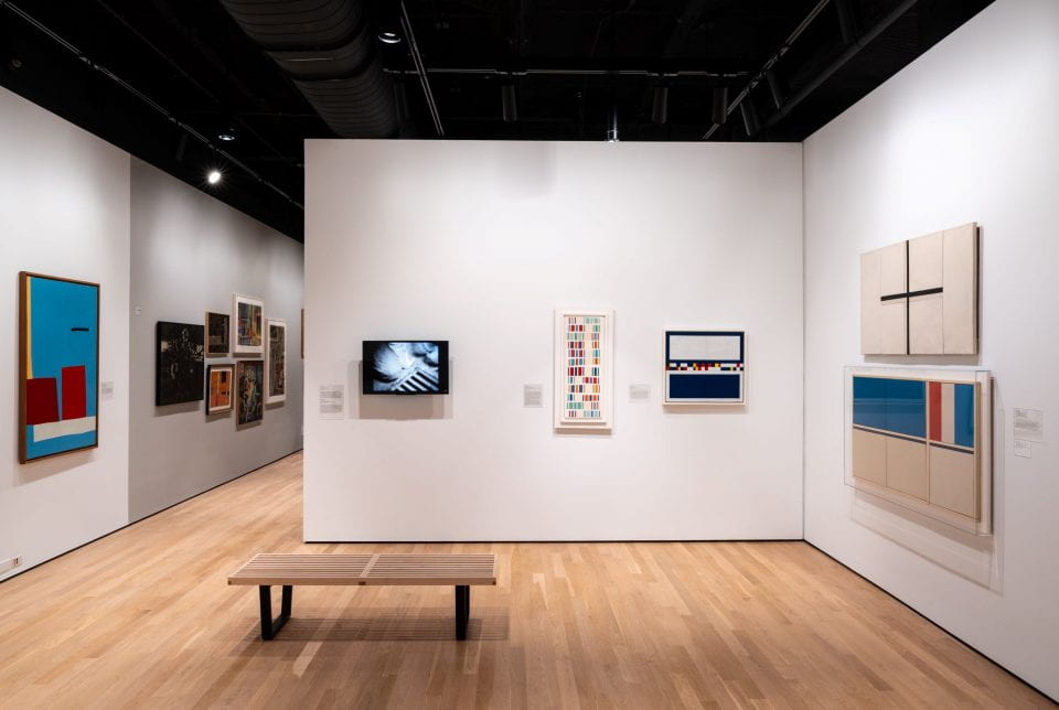 An image of a gallery wall with a video monitor. There is an empty bench in front of it.