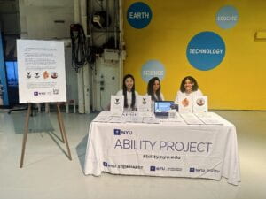 At the center is a table covered with a white cloth bearing the NYU Ability Project. On the table, there are pictures of patches and digitally embroidered patches as well as Swell form patches. There is a laptop displaying a video of the digital embroidery machine in operation. There are 3 women sitting at the table and wearing white tee shirts which say "NYU Ability Project." There is a yellow wall in the background with blue circles and words inside which say, "Earth", "Educate", "Science", and "Technology." There is a poster on an easel describing the purpose of digitally embroidered patches.