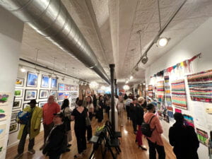 Photo of the inside of Clover's Fine Art Gallery on the opening day of Artshare's gallery exhibition. Room is filled with visitors and artwork, including paintings, tactile pieces, and drawings which are hung up on all the walls. 