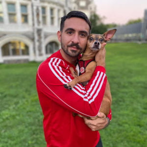 Dave Whitman is wearing a red tracksuit and holding a small dog up to his face. They are standing on a lawn in front of a house. The background is blurred.