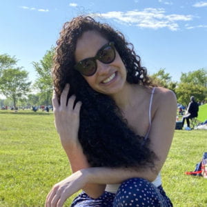 Dannysha Diaz smiling in a park. She is wearing sunglasses and tucking her hair behind her ear.