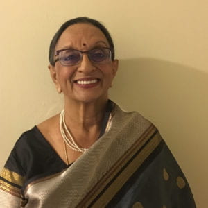 Vasu Varadhan smiling. She is standing against a plain background and is wearing a sari and a pearl necklace.