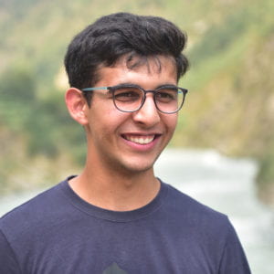 Moosa Waraich smiling. He is standing in front of a blurred background of trees and is wearing glasses.