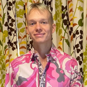 Van Tingley stands in front of a floral patterned wallpaper. wearing a pink patterned shirt. The shirt and wallpaper are clashing.