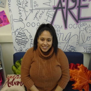 Jasmine Stakley smiles up at the camera. She is seated in her classroom in front of a wall of posters, and wears a turtleneck sweater and ponytail.