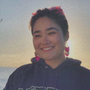 Charlie Dodge smiling. She is standing in front of a sunset and is wearing a sweatshirt, dangling earrings, and pink sunglasses on top of her head.