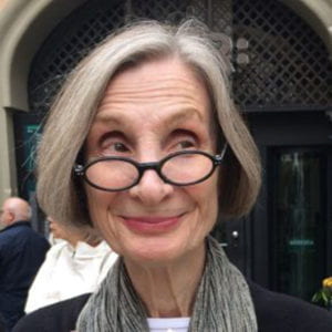 June Foley smiling. She is looking off to the side and her oval glasses are pushed down her nose.