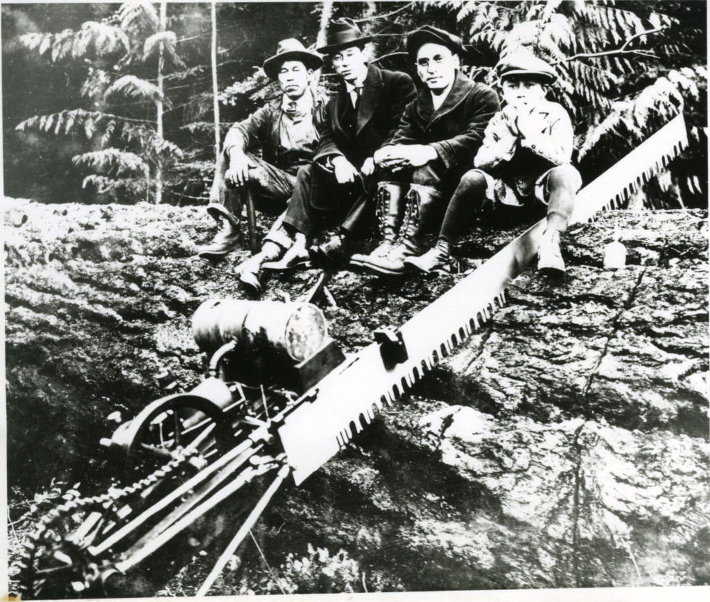 Black and white portrait of three men and a boy standing in front of a large felled log with an axe and small dog resting on the tree stump; the men wear boots, caps or hats; location identified as Island Center, Bainbridge Island, Washington and individuals as Mr. Harui and friends.