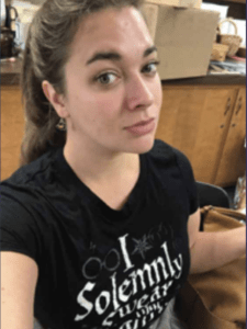 Color portrait of Alexsandra Gizzi in front of a wooden cabinet, wearing a black graphic tee with white lettering.