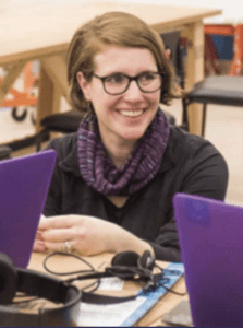 Color portrait of Amy Hurst at a desk with laptops, wearing glasses, a dark grey sweater with multicolor scarf.