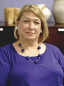 Color portrait of Beth Ziebarth in front of a wood shelf with various vases on top, wearing a purple shirt and matching necklace.