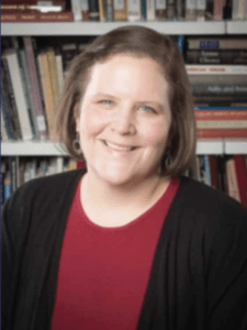 Color portrait of Carrie Villar in front of a bookshelf, wearing a red shirt with black cardigan.