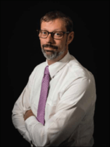 Color portrait of Chris Malanson on black background, wearing glasses, a white collared shirt and pink tie.