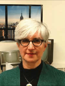 Color portrait of Elaine Charnov in front of a picture of NYC skyline, wearing glasses, a green sweater and black shirt.