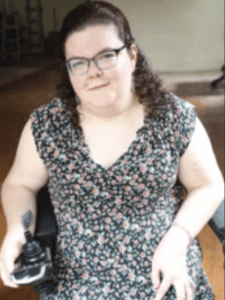 Color portrait of Emily Ladau in front of a wood background and white wall, wearing glasses and a floral dress.