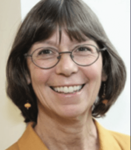 Color portrait of Janet Rassweiler on a white background, wearing glasses, a mustard yellow shirt and gold pendant earrings.