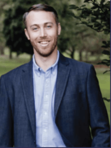 Color portrait of Kyle Sheperd in front of a lawn with tall trees, wearing a light blue button down shirt and navy blazer.