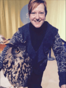 Color portrait of Lynda Kennedy on a yellow background, wearing a blue sweater with black turtleneck. There is a black and white horned-owl in the foreground.