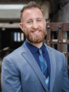 Color portrait of Matthew Murphy in front of a metal grate, wearing a blue button down, multicolor blue tie and blue blazer.