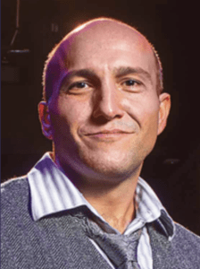 Color portrait of Michael Tranquilli on a black background, wearing a white and grey striped button down, grey tie and grey sweater.