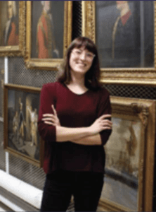 Color portrait of Miranda Peters in front of five frame paintings, wearing glasses, a maroon shirt and black pants.