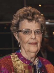 Color portrait of Ruth Bernstein on a black background, wearing glasses and a multicolor jacket.