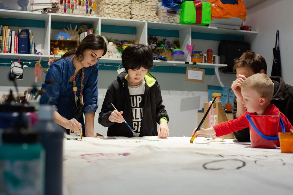 adults and children sit around a table painting on a large canvas