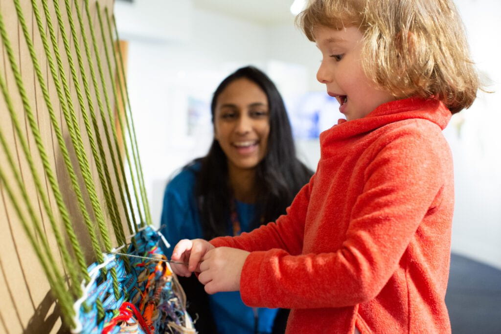 A child is weaving string and smiling while an adult looks on in the background