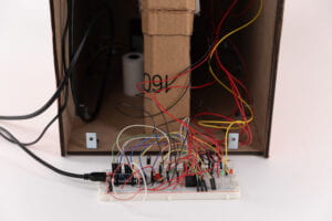 close up View of the back and internal components of "Bon Appetit" on the game console including thermal printer, arduino, wires attached to breadboard, and cardboard dispenser